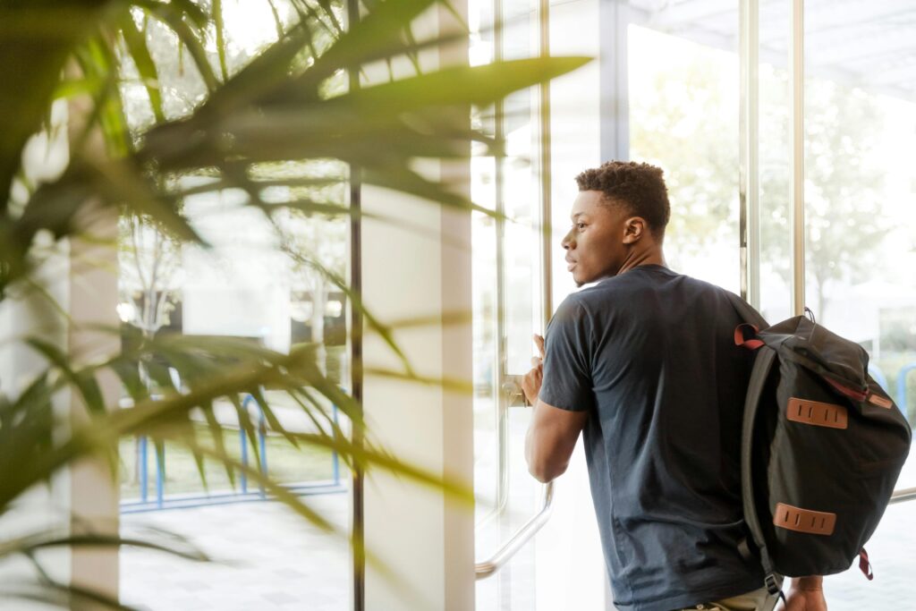 A college student with a backpack exits a campus building through glass doors.